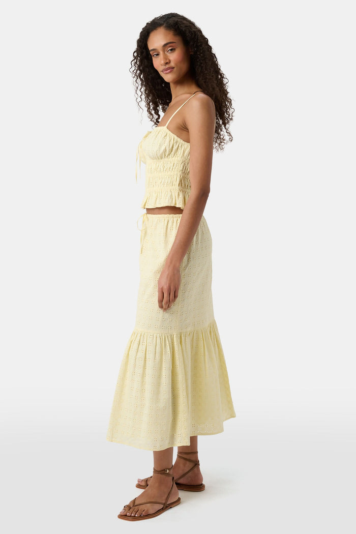 A woman with curly hair wears the Roller Rabbit Petal Eyelet Aurelia Top, a yellow cotton camisole with eyelet details and a ruffled hem, paired with a matching high-waisted skirt against a plain light background.