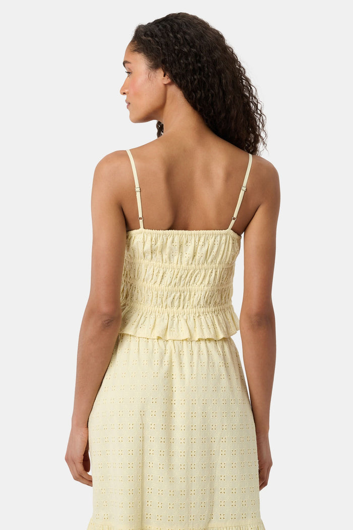 A woman with curly hair wears the Roller Rabbit Petal Eyelet Aurelia Top, a yellow cotton camisole with eyelet details and a ruffled hem, paired with a matching high-waisted skirt against a plain light background.