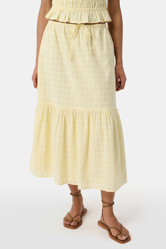 A person wears the Roller Rabbit Petal Eyelet Goldie Skirt, a butter yellow midi with eyelet details, ruffled hem, and gathered top, paired with strappy brown sandals. Their hands rest relaxed by their sides.