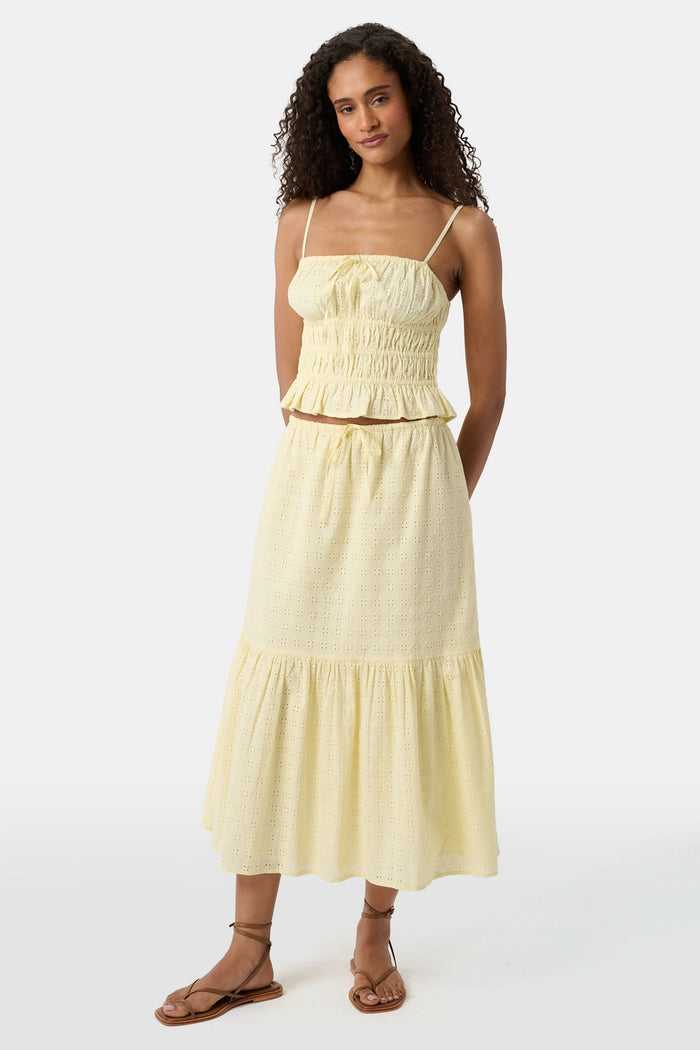 A woman with curly hair wears the Roller Rabbit Petal Eyelet Aurelia Top, a yellow cotton camisole with eyelet details and a ruffled hem, paired with a matching high-waisted skirt against a plain light background.