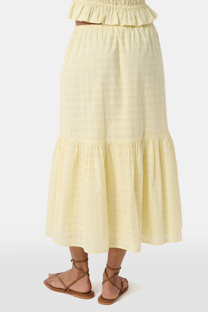 A person wears the Roller Rabbit Petal Eyelet Goldie Skirt, a butter yellow midi with eyelet details, ruffled hem, and gathered top, paired with strappy brown sandals. Their hands rest relaxed by their sides.
