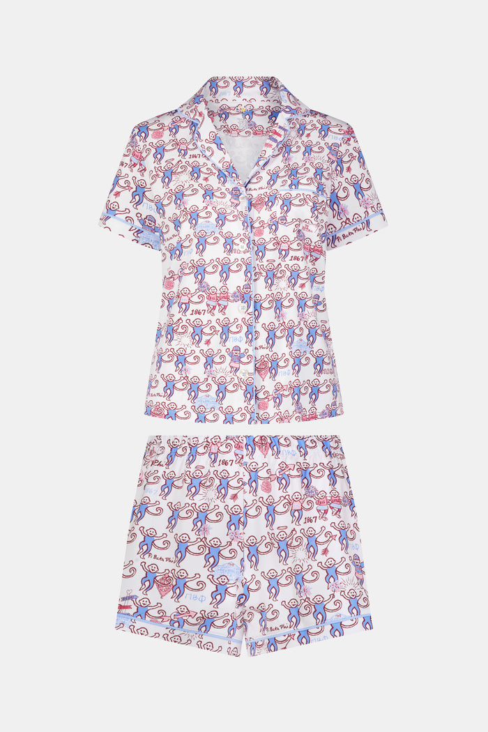 The Roller Rabbit Pi Beta Phi Monkey Polo Pajamas feature a short-sleeved button-up top and matching shorts, adorned with a playful pink and blue monkey print on a white background. Crafted from soft Pima cotton with an elastic waistband for comfort, these pajamas are made in Peru.