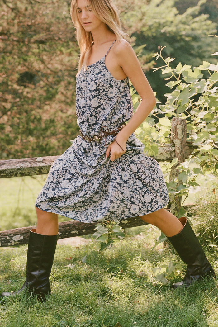 A woman in the Roller Rabbit Priya Ditsy Augustine Dress and black boots stands outdoors by a wooden fence with green foliage and trees in the background.