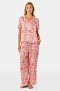 A woman stands barefoot against a plain white background, wearing the Roller Rabbit Priya Floral Alvie Silk Set—a matching pink floral pajama ensemble featuring short sleeves and long pants, with one hand in her pocket. This washable silk loungewear set offers both comfort and style.