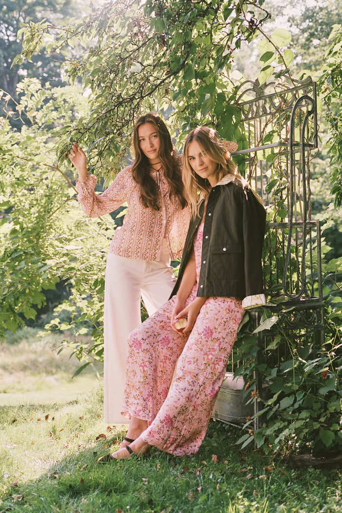 A woman stands barefoot against a plain white background, wearing the Roller Rabbit Priya Floral Alvie Silk Set—a matching pink floral pajama ensemble featuring short sleeves and long pants, with one hand in her pocket. This washable silk loungewear set offers both comfort and style.