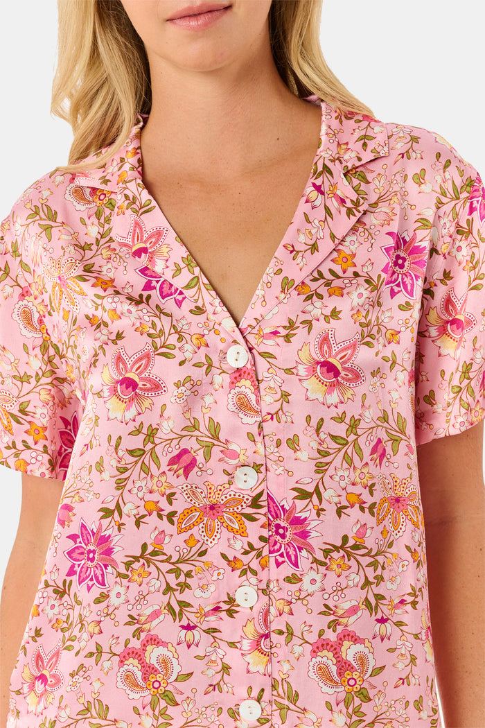 A woman stands barefoot against a plain white background, wearing the Roller Rabbit Priya Floral Alvie Silk Set—a matching pink floral pajama ensemble featuring short sleeves and long pants, with one hand in her pocket. This washable silk loungewear set offers both comfort and style.