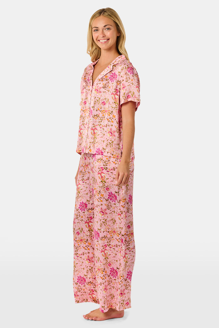 A woman stands barefoot against a plain white background, wearing the Roller Rabbit Priya Floral Alvie Silk Set—a matching pink floral pajama ensemble featuring short sleeves and long pants, with one hand in her pocket. This washable silk loungewear set offers both comfort and style.