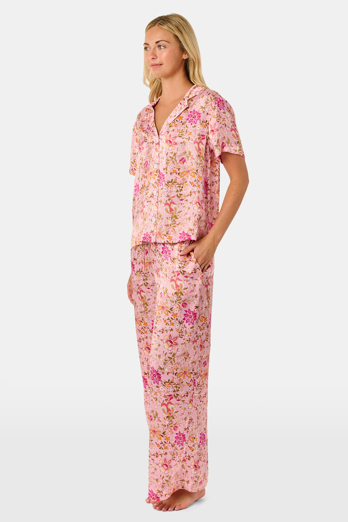 A woman stands barefoot against a plain white background, wearing the Roller Rabbit Priya Floral Alvie Silk Set—a matching pink floral pajama ensemble featuring short sleeves and long pants, with one hand in her pocket. This washable silk loungewear set offers both comfort and style.