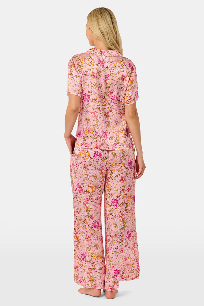 A woman stands barefoot against a plain white background, wearing the Roller Rabbit Priya Floral Alvie Silk Set—a matching pink floral pajama ensemble featuring short sleeves and long pants, with one hand in her pocket. This washable silk loungewear set offers both comfort and style.