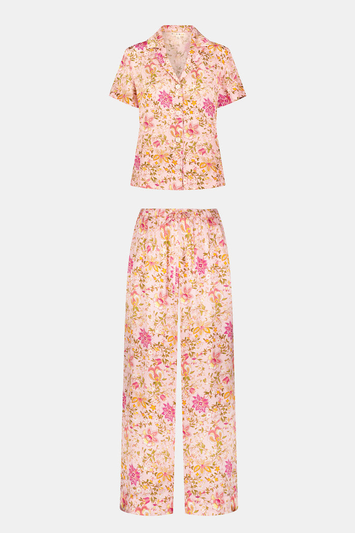 A woman stands barefoot against a plain white background, wearing the Roller Rabbit Priya Floral Alvie Silk Set—a matching pink floral pajama ensemble featuring short sleeves and long pants, with one hand in her pocket. This washable silk loungewear set offers both comfort and style.