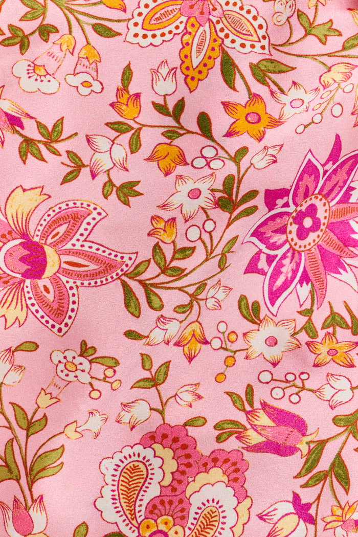 A woman stands barefoot against a plain white background, wearing the Roller Rabbit Priya Floral Alvie Silk Set—a matching pink floral pajama ensemble featuring short sleeves and long pants, with one hand in her pocket. This washable silk loungewear set offers both comfort and style.