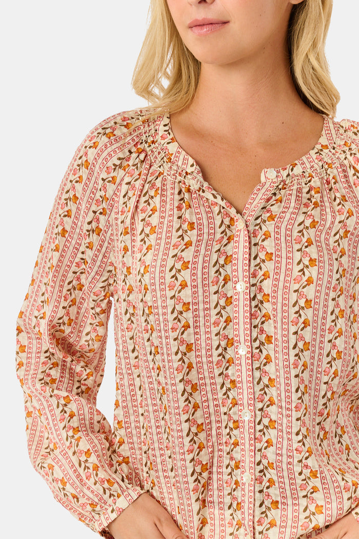 A woman stands smiling with one hand in her pocket against a plain white background, wearing the Roller Rabbit Priya Floral Colleen Top.