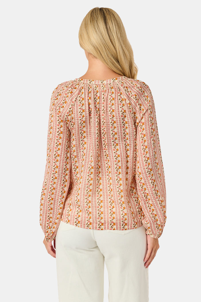 A woman stands smiling with one hand in her pocket against a plain white background, wearing the Roller Rabbit Priya Floral Colleen Top.