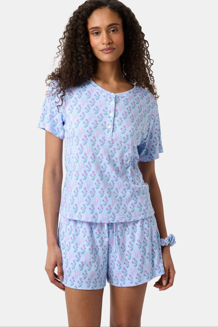 A woman with long curly hair wears the Rosa Ditsy Monkey Alexa Set by Roller Rabbit, a light blue Pima cotton pajama set featuring a ditsy monkey print. The set includes a henley-style top and matching shorts. She stands against a plain background.