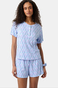 A woman with long curly hair wears the Rosa Ditsy Monkey Alexa Set by Roller Rabbit, a light blue Pima cotton pajama set featuring a ditsy monkey print. The set includes a henley-style top and matching shorts. She stands against a plain background.