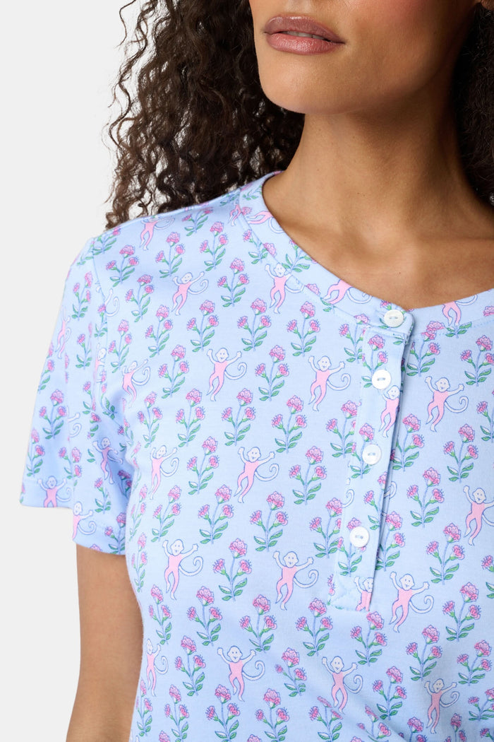 A woman with long curly hair wears the Rosa Ditsy Monkey Alexa Set by Roller Rabbit, a light blue Pima cotton pajama set featuring a ditsy monkey print. The set includes a henley-style top and matching shorts. She stands against a plain background.