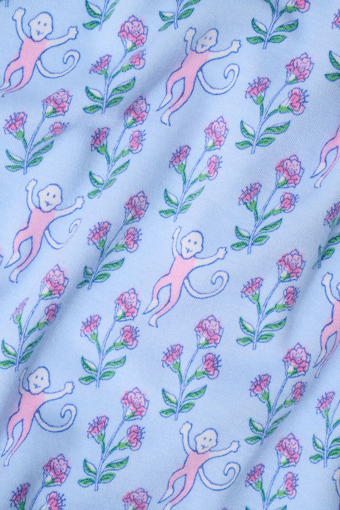 A woman with long curly hair wears the Rosa Ditsy Monkey Alexa Set by Roller Rabbit, a light blue Pima cotton pajama set featuring a ditsy monkey print. The set includes a henley-style top and matching shorts. She stands against a plain background.