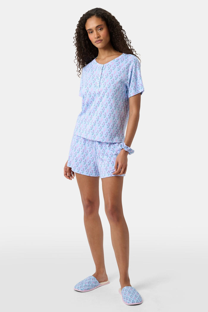 A woman with long curly hair wears the Rosa Ditsy Monkey Alexa Set by Roller Rabbit, a light blue Pima cotton pajama set featuring a ditsy monkey print. The set includes a henley-style top and matching shorts. She stands against a plain background.