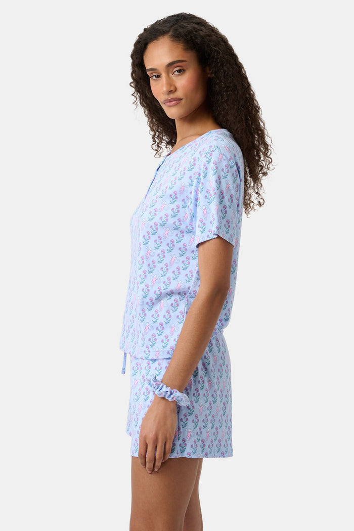 A woman with long curly hair wears the Rosa Ditsy Monkey Alexa Set by Roller Rabbit, a light blue Pima cotton pajama set featuring a ditsy monkey print. The set includes a henley-style top and matching shorts. She stands against a plain background.