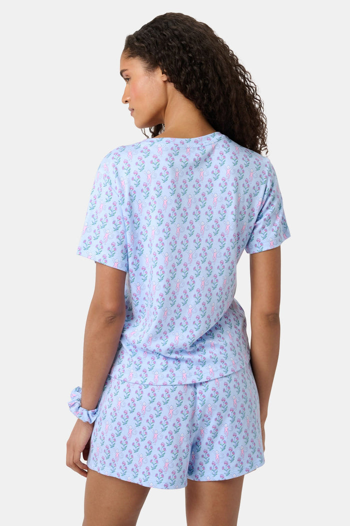 A woman with long curly hair wears the Rosa Ditsy Monkey Alexa Set by Roller Rabbit, a light blue Pima cotton pajama set featuring a ditsy monkey print. The set includes a henley-style top and matching shorts. She stands against a plain background.