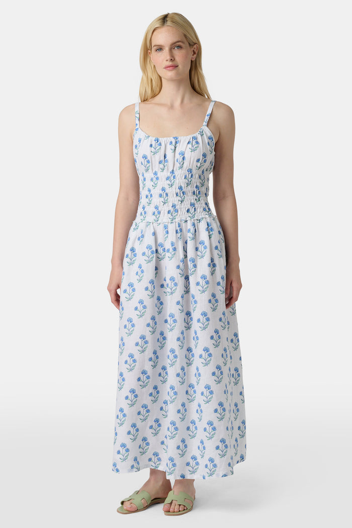 A woman wears the Roller Rabbit Rosa Floral Veronica Dress, a white sundress with blue prints, a smocked bodice, and spaghetti straps, styled with light green sandals as she stands facing forward with her arms relaxed by her sides.