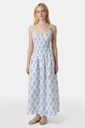 A woman wears the Roller Rabbit Rosa Floral Veronica Dress, a white sundress with blue prints, a smocked bodice, and spaghetti straps, styled with light green sandals as she stands facing forward with her arms relaxed by her sides.