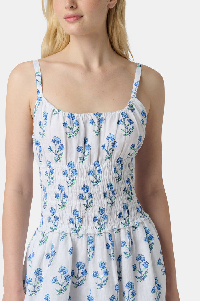 A woman wears the Roller Rabbit Rosa Floral Veronica Dress, a white sundress with blue prints, a smocked bodice, and spaghetti straps, styled with light green sandals as she stands facing forward with her arms relaxed by her sides.
