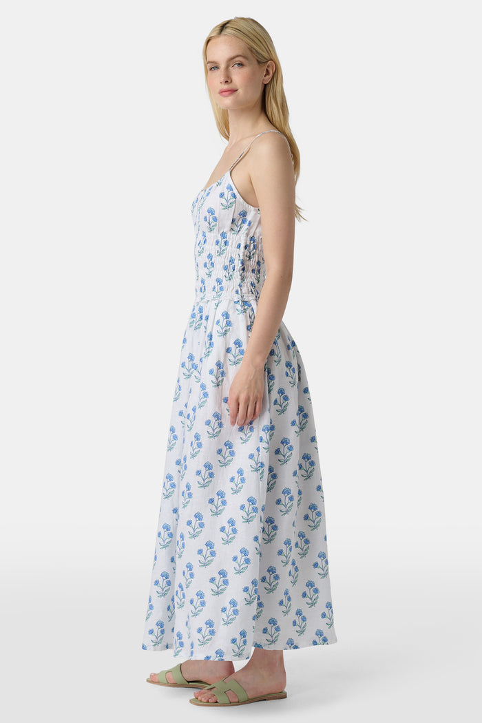 A woman wears the Roller Rabbit Rosa Floral Veronica Dress, a white sundress with blue prints, a smocked bodice, and spaghetti straps, styled with light green sandals as she stands facing forward with her arms relaxed by her sides.
