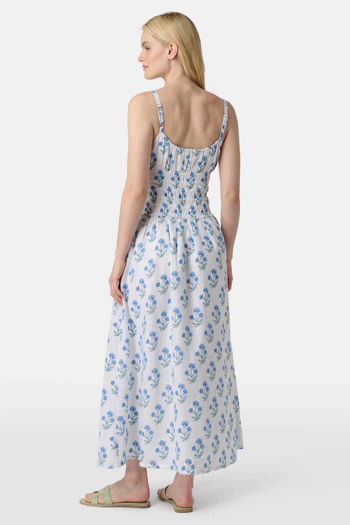 A woman wears the Roller Rabbit Rosa Floral Veronica Dress, a white sundress with blue prints, a smocked bodice, and spaghetti straps, styled with light green sandals as she stands facing forward with her arms relaxed by her sides.