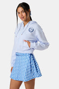 A woman wears the Roller Rabbit RR Athletic Club Cropped Zip Up—a pastel blue terry hoodie with an athletic club logo on the chest—paired with a blue-and-white checkered skirt, standing against a white background.