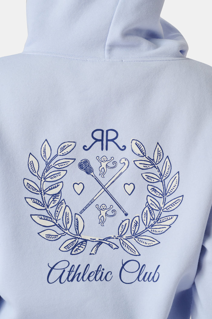 A woman wears the Roller Rabbit RR Athletic Club Cropped Zip Up—a pastel blue terry hoodie with an athletic club logo on the chest—paired with a blue-and-white checkered skirt, standing against a white background.