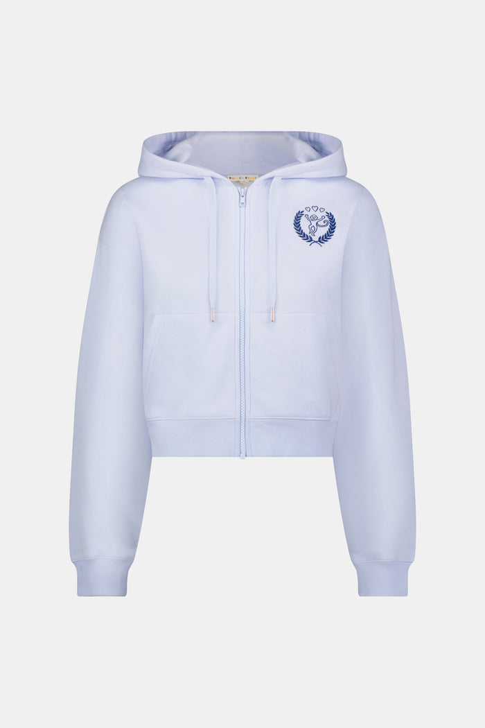 A woman wears the Roller Rabbit RR Athletic Club Cropped Zip Up—a pastel blue terry hoodie with an athletic club logo on the chest—paired with a blue-and-white checkered skirt, standing against a white background.