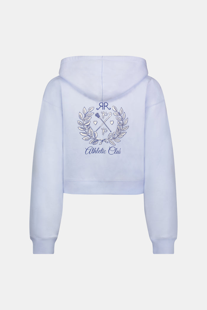 A woman wears the Roller Rabbit RR Athletic Club Cropped Zip Up—a pastel blue terry hoodie with an athletic club logo on the chest—paired with a blue-and-white checkered skirt, standing against a white background.
