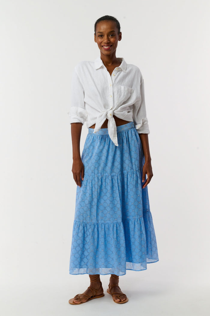 A person in a white, long-sleeved button-up shirt tied at the waist, the Roller Rabbit Ruban Carlisa Skirt, and brown strappy sandals stands against a white background.