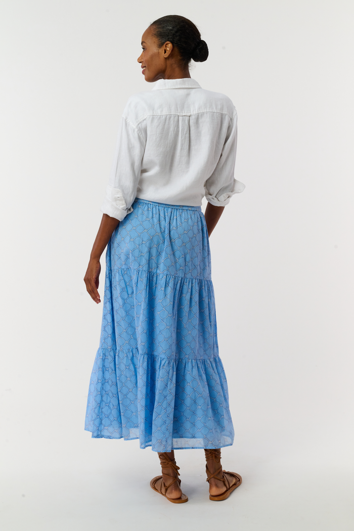 A person in a white, long-sleeved button-up shirt tied at the waist, the Roller Rabbit Ruban Carlisa Skirt, and brown strappy sandals stands against a white background.