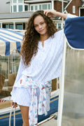 A woman with long curly hair smiles softly by a blue boat with striped canopy, wearing the Seaside Knots Ella Set by Roller Rabbit in a relaxed outdoor setting.