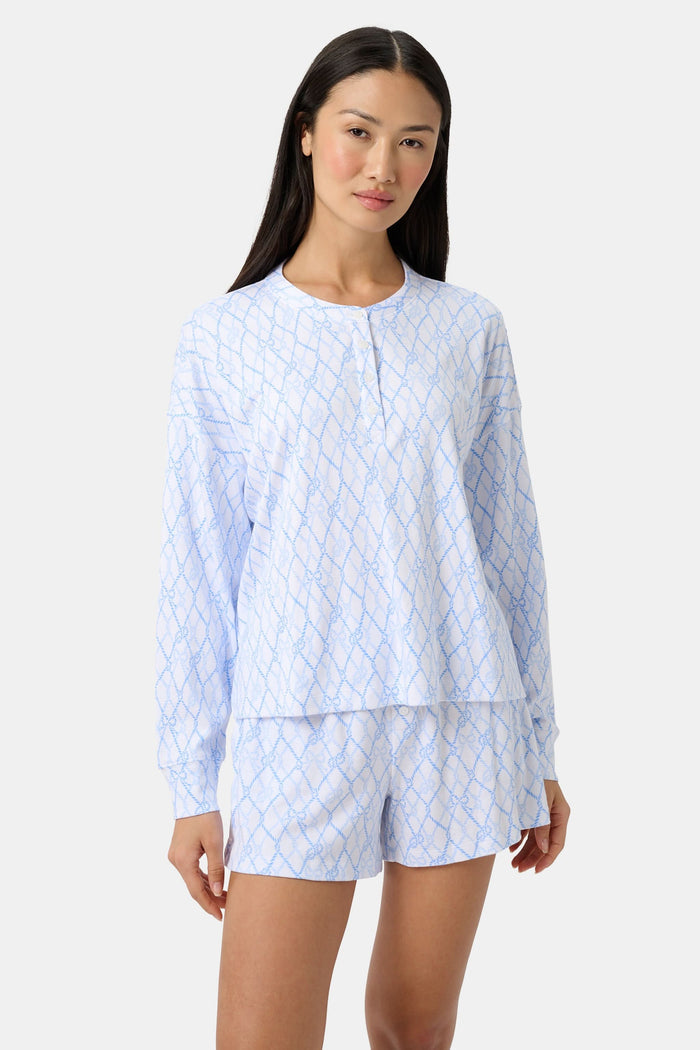 A woman with long curly hair smiles softly by a blue boat with striped canopy, wearing the Seaside Knots Ella Set by Roller Rabbit in a relaxed outdoor setting.