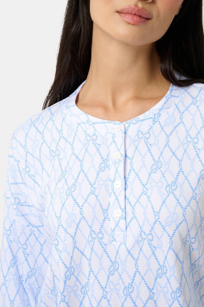 A woman with long curly hair smiles softly by a blue boat with striped canopy, wearing the Seaside Knots Ella Set by Roller Rabbit in a relaxed outdoor setting.