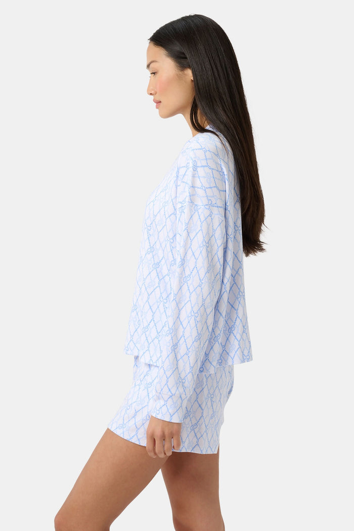 A woman with long curly hair smiles softly by a blue boat with striped canopy, wearing the Seaside Knots Ella Set by Roller Rabbit in a relaxed outdoor setting.