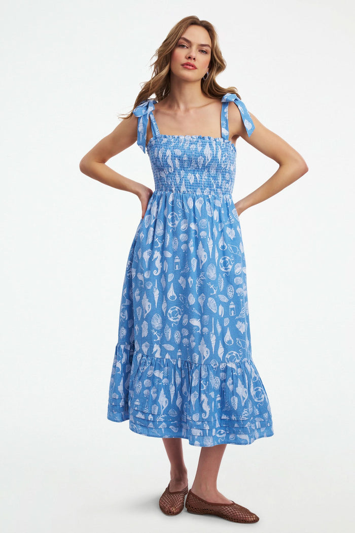 A woman wears the Roller Rabbit Seaside Stripe Georgina Dress, featuring light blue stripes, a smocked bodice, ruffled hem, and tie straps. She stands with one leg bent and pairs the look with brown woven flats.