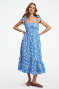 A woman wears the Roller Rabbit Seaside Stripe Georgina Dress, featuring light blue stripes, a smocked bodice, ruffled hem, and tie straps. She stands with one leg bent and pairs the look with brown woven flats.
