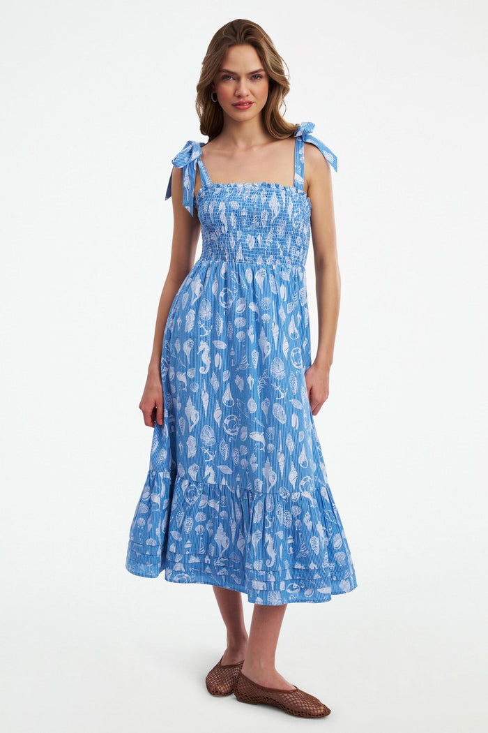 A woman wears the Roller Rabbit Seaside Stripe Georgina Dress, featuring light blue stripes, a smocked bodice, ruffled hem, and tie straps. She stands with one leg bent and pairs the look with brown woven flats.