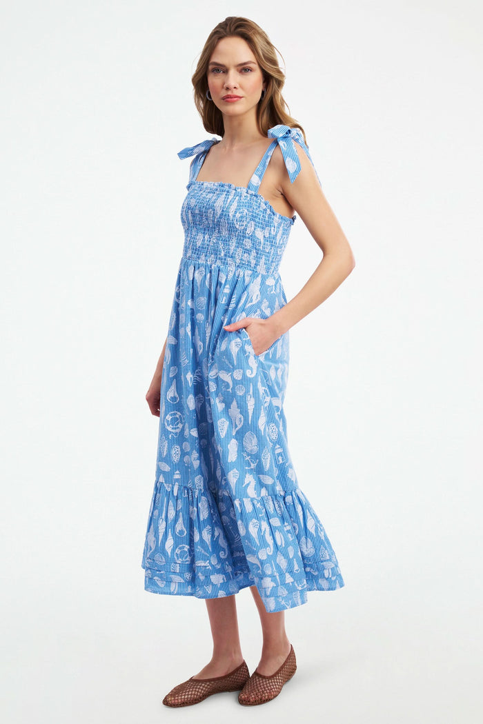 A woman wears the Roller Rabbit Seaside Stripe Georgina Dress, featuring light blue stripes, a smocked bodice, ruffled hem, and tie straps. She stands with one leg bent and pairs the look with brown woven flats.