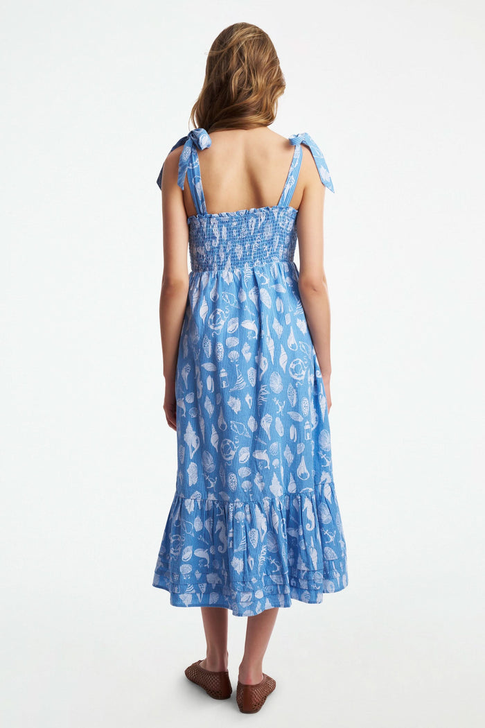 A woman wears the Roller Rabbit Seaside Stripe Georgina Dress, featuring light blue stripes, a smocked bodice, ruffled hem, and tie straps. She stands with one leg bent and pairs the look with brown woven flats.