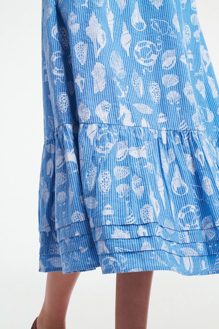 A woman wears the Roller Rabbit Seaside Stripe Georgina Dress, featuring light blue stripes, a smocked bodice, ruffled hem, and tie straps. She stands with one leg bent and pairs the look with brown woven flats.