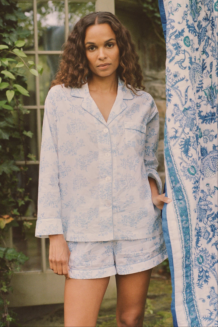 A woman with curly hair stands outdoors near a glass door and patterned fabric, wearing the Roller Rabbit Sherwood Forest Monkey Leila Lounge Set, with greenery in the background.