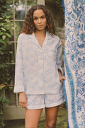 A woman with curly hair stands outdoors near a glass door and patterned fabric, wearing the Roller Rabbit Sherwood Forest Monkey Leila Lounge Set, with greenery in the background.