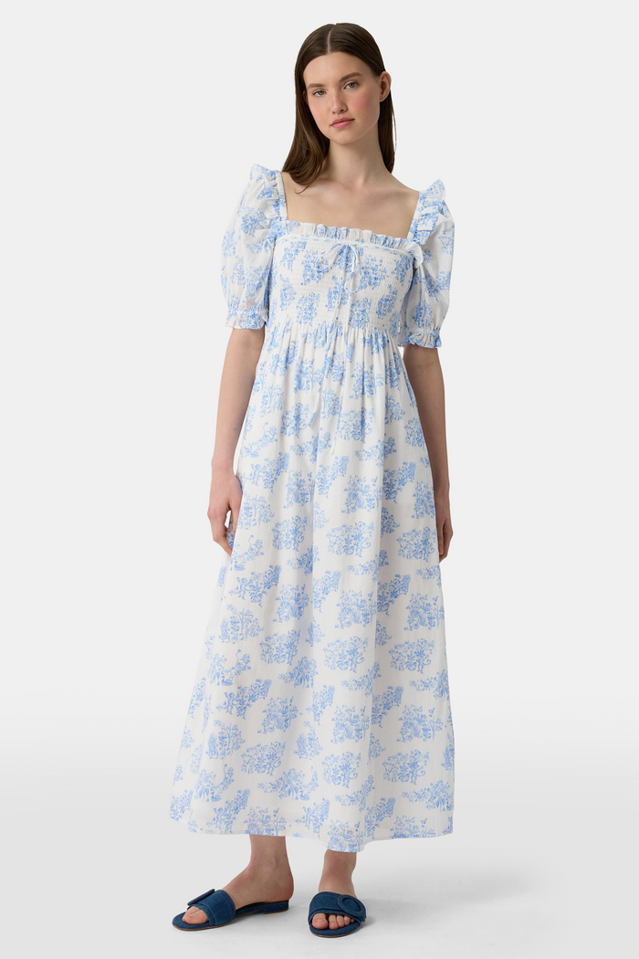 A woman wears the Roller Rabbit Sherwood Forest Monkey Scarlett Nap Dress—long, white with a blue monkey toile print, puffed short sleeves, smocked bodice, and square neckline—paired with blue slip-on sandals against a light background.