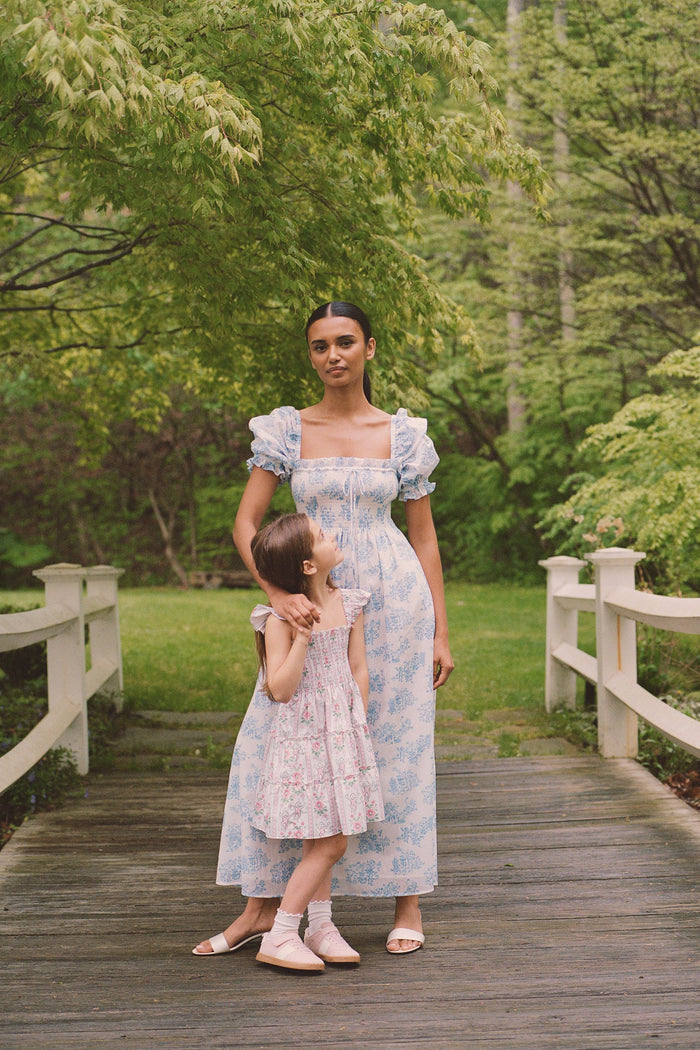 A woman wears the Roller Rabbit Sherwood Forest Monkey Scarlett Nap Dress—long, white with a blue monkey toile print, puffed short sleeves, smocked bodice, and square neckline—paired with blue slip-on sandals against a light background.
