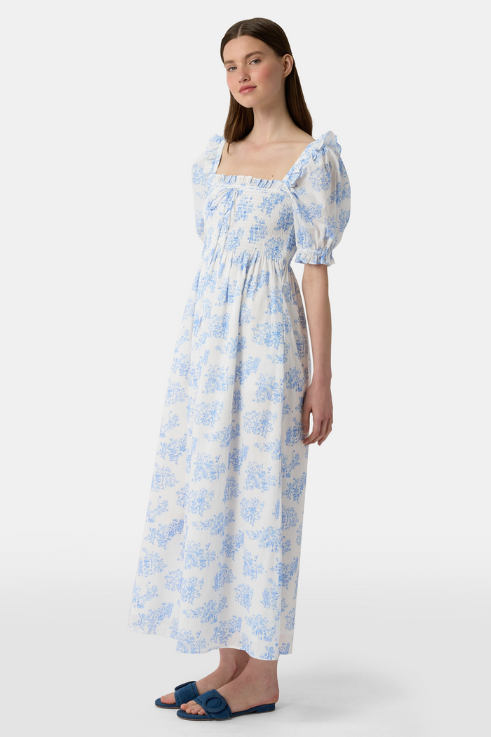 A woman wears the Roller Rabbit Sherwood Forest Monkey Scarlett Nap Dress—long, white with a blue monkey toile print, puffed short sleeves, smocked bodice, and square neckline—paired with blue slip-on sandals against a light background.